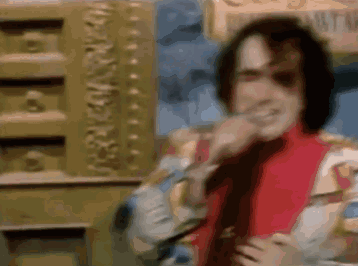 A Man In A Red Jacket And Tie Is Standing In Front Of A Gold Wall And Smiling . GIF