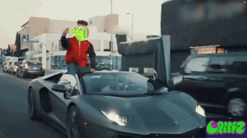A Man In A Red Jacket Is Driving A Lamborghini With A Frog Face On The Hood GIF