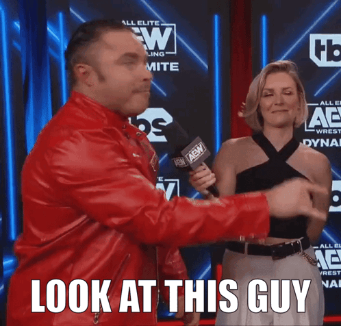 A Man In A Red Jacket Is Holding A Microphone And A Woman Behind Him Says " Look At This Guy " GIF