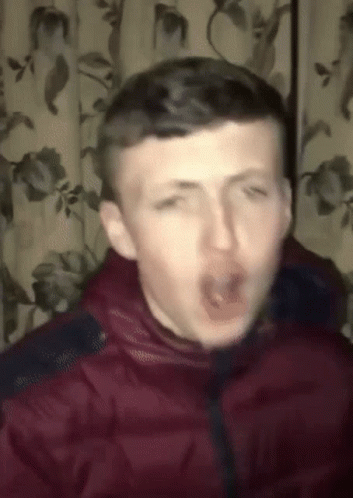 A Man In A Red Jacket Is Making A Funny Face In Front Of A Curtain With Flowers On It GIF