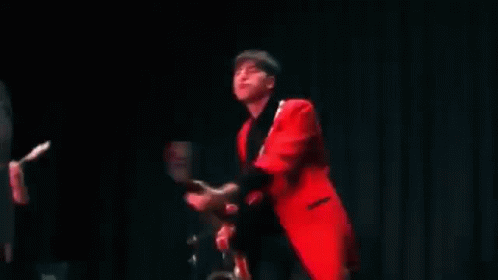 A Man In A Red Jacket Is Playing A Guitar In A Band . GIF