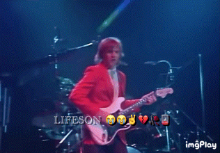 A Man In A Red Jacket Is Playing A Guitar On A Stage With The Words Lifeson On The Bottom Right GIF