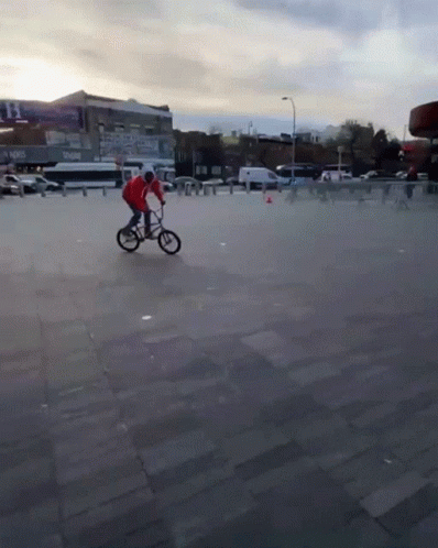 A Man In A Red Jacket Is Riding A Bike On A Concrete Pavement GIF