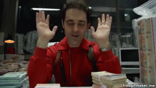 A Man In A Red Jacket Is Sitting At A Desk With A Pile Of Money . GIF