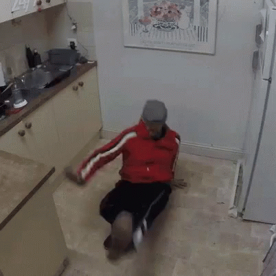 A Man In A Red Jacket Is Sitting On The Floor In A Kitchen With A Picture On The Wall Above Him . GIF