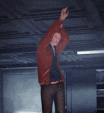 A Man In A Red Jacket Is Standing In A Dark Room With His Hands In The Air GIF