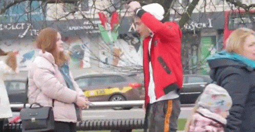 A Man In A Red Jacket Is Standing Next To A Woman In A Pink Jacket On A City Street . GIF