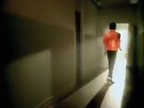 A Man In A Red Jacket Is Walking Down A Dark Hallway Towards A Light . GIF