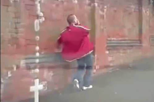 A Man In A Red Jacket Is Walking In Front Of A Red Brick Wall . GIF