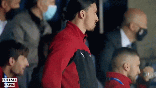 A Man In A Red Jacket Stands In A Crowd With The Word Futbol Karim On The Bottom Left GIF