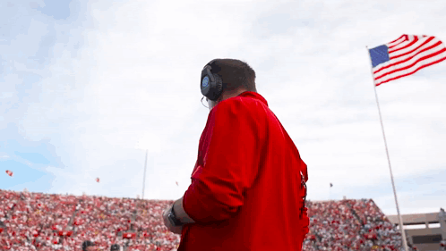 A Man In A Red Jacket Stands In Front Of A Large American Flag GIF