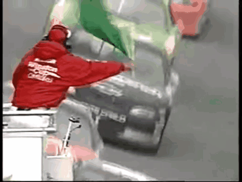 A Man In A Red Jacket That Says Windstar On It Stands Next To A Race Car GIF