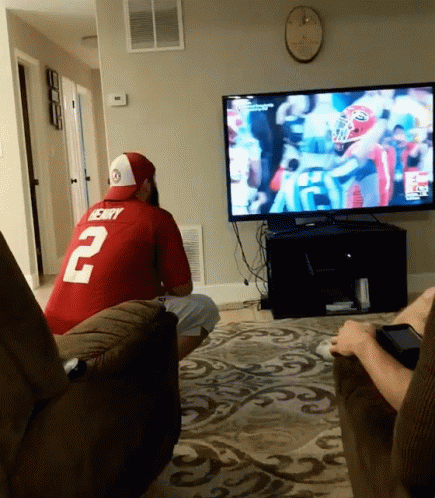A Man In A Red Jersey With The Number 2 On It Sits In Front Of A Tv GIF