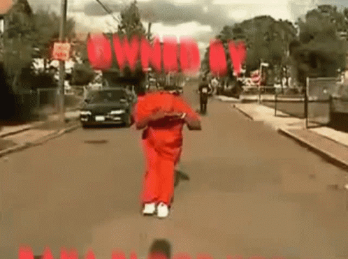 A Man In A Red Jumpsuit Is Running Down A Street With A Car Behind Him . GIF