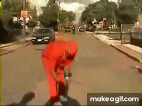A Man In A Red Jumpsuit Is Standing On A Street . GIF
