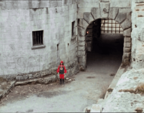 A Man In A Red Knight Costume Stands In Front Of A Stone Building GIF