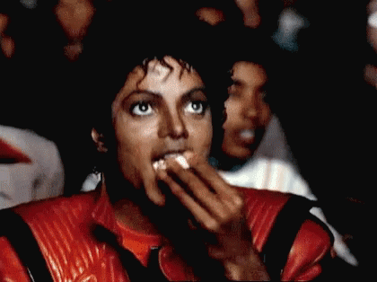 A Man In A Red Leather Jacket Is Covering His Mouth With His Hands While Watching A Movie . GIF