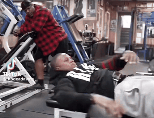 A Man In A Red Plaid Shirt Is Standing Next To A Man In A Black And White Shirt On A Leg Press Machine . GIF
