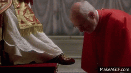 A Man In A Red Robe Is Kissing The Feet Of A Man In A White Dress . GIF