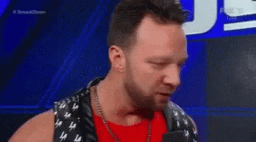 A Man In A Red Shirt And Black Vest Is Talking Into A Microphone On A Stage . GIF