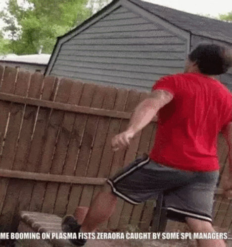 A Man In A Red Shirt And Gray Shorts Is Jumping Over A Wooden Fence In Front Of A House . GIF