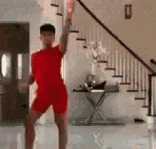 A Man In A Red Shirt And Shorts Is Dancing In A Living Room Next To Stairs . GIF