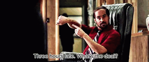 A Man In A Red Shirt And Tie Is Sitting In A Chair And Says Three Hours Late What 'S The Deal GIF