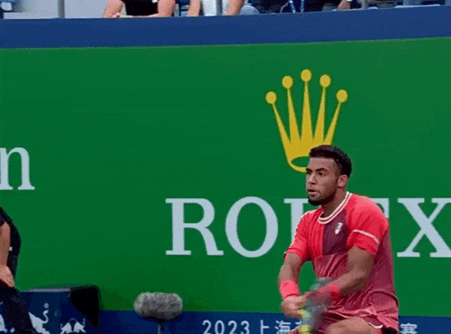 A Man In A Red Shirt Holds A Tennis Racquet In Front Of A Rolex Sign GIF