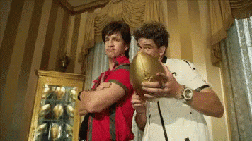 A Man In A Red Shirt Is Holding A Football Next To Another Man In A White Shirt GIF