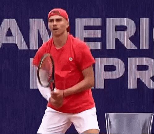 A Man In A Red Shirt Is Holding A Tennis Racket In Front Of A Blue Background . GIF