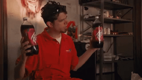 A Man In A Red Shirt Is Holding Two Bottles Of Soda One Of Which Has The Letter A On It GIF
