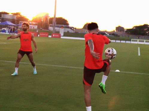 A Man In A Red Shirt Is Kicking A Soccer Ball While Another Man Looks On GIF
