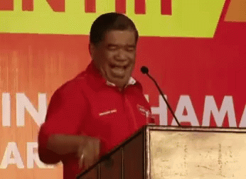 A Man In A Red Shirt Is Laughing While Standing At A Podium With The Word Ham On The Wall Behind Him GIF