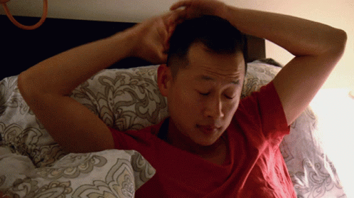 A Man In A Red Shirt Is Laying In Bed With His Hands On His Head GIF