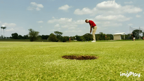 A Man In A Red Shirt Is Putting A Ball Into A Hole On A Golf Course GIF
