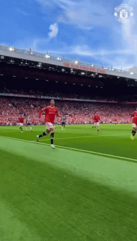 A Man In A Red Shirt Is Running On A Soccer Field In Front Of A Manchester United Stadium GIF