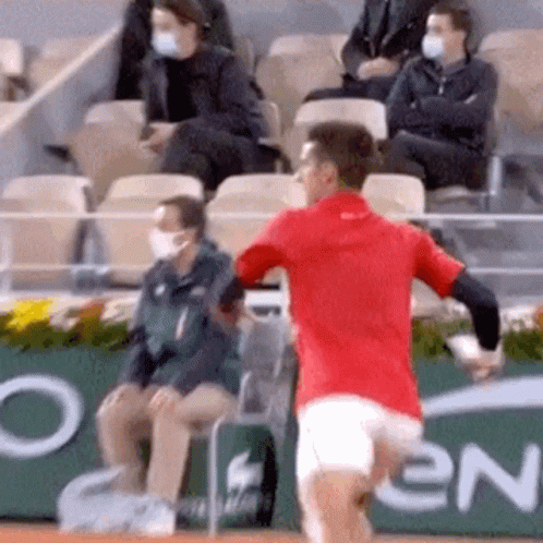 A Man In A Red Shirt Is Running On A Tennis Court In Front Of A Crowd Wearing Masks . GIF