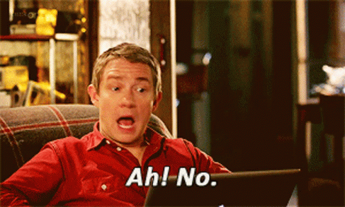 A Man In A Red Shirt Is Sitting On A Couch Looking At A Laptop And Saying Ah No GIF