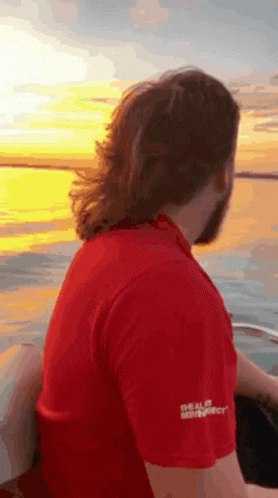 A Man In A Red Shirt Is Sitting On The Back Of A Boat Looking At The Sunset . GIF