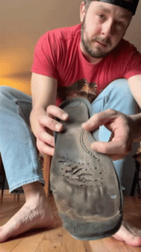 A Man In A Red Shirt Is Sitting On The Floor Holding A Shoe Sole GIF
