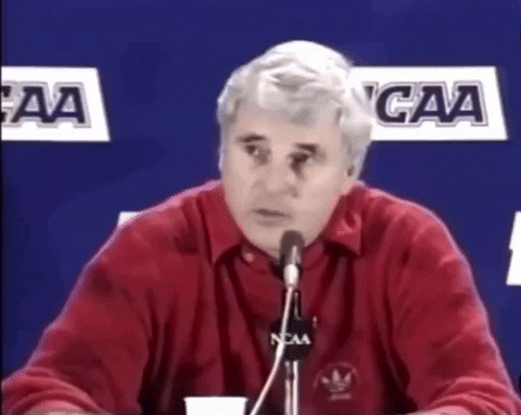 A Man In A Red Shirt Is Speaking Into A Microphone With Ncaa Logos In The Background GIF