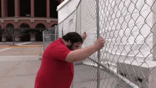 A Man In A Red Shirt Is Standing Behind A Chain Link Fence GIF