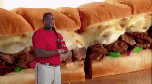 A Man In A Red Shirt Is Standing In Front Of A Very Large Sandwich . GIF