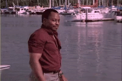 A Man In A Red Shirt Is Standing Next To A Body Of Water With Boats In The Background . GIF
