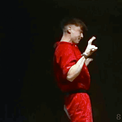 A Man In A Red Shirt Is Standing On A Stage With His Hands Up GIF