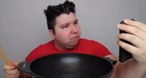 a man in a red shirt is taking a picture of a wok with a phone