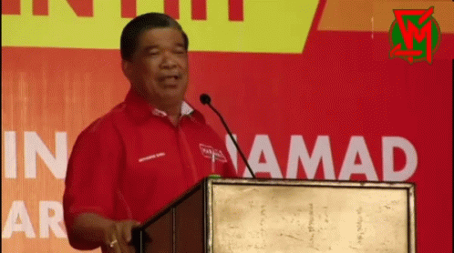 A Man In A Red Shirt Stands At A Podium In Front Of A Sign That Says In Amada GIF