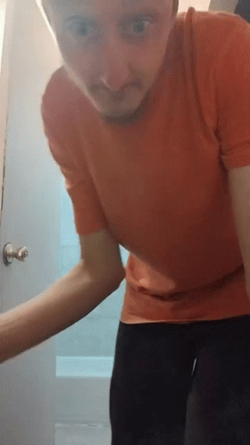 A Man In A Red Shirt Stands In Front Of A Door GIF
