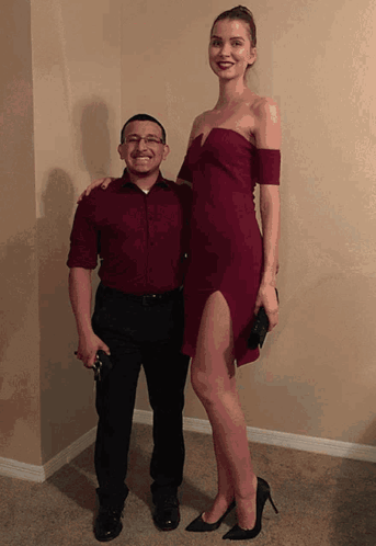 A Man In A Red Shirt Stands Next To A Very Tall Woman In A Red Dress GIF