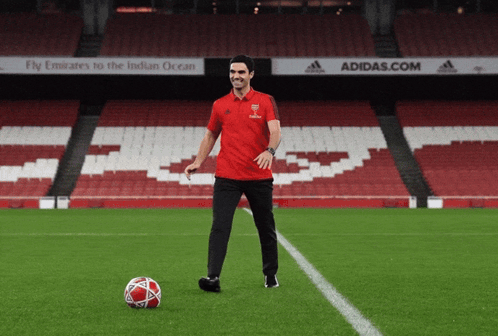 A Man In A Red Shirt Stands On A Soccer Field In Front Of A Sign That Says Adidas.com GIF